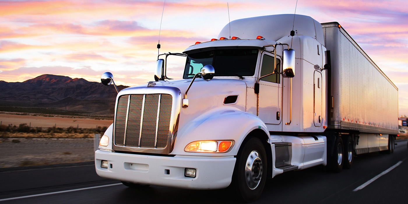 White semi-truck pulling a long trailer driving on a highway with mountains and a colorful sunset sky in the background