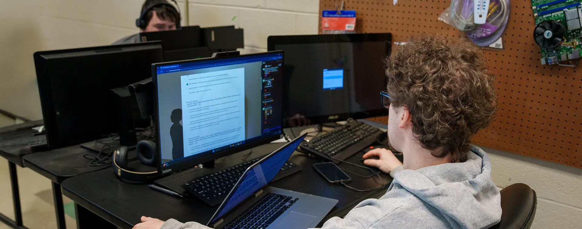 Student using dual monitors and laptop at desk with headset nearby; document open on screen in computer lab