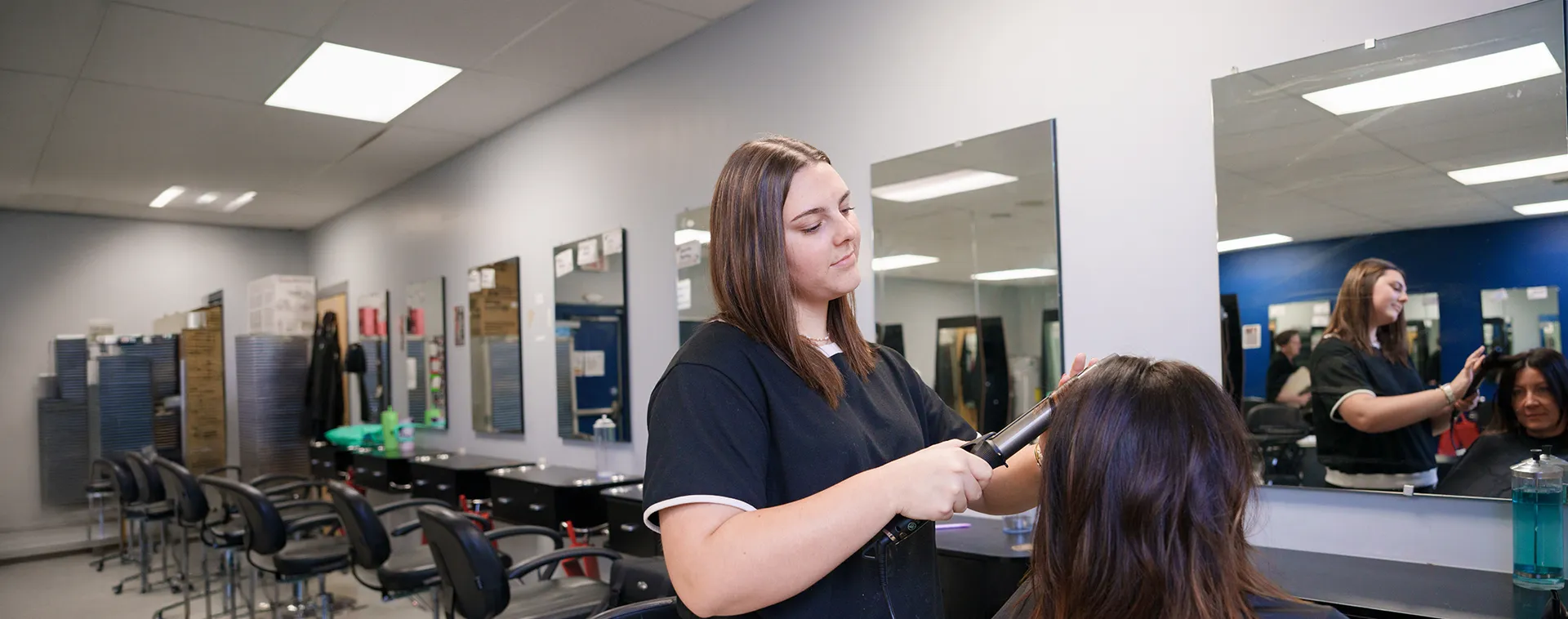 Student styling hair with curling iron in salon classroom with mirrors and chairs