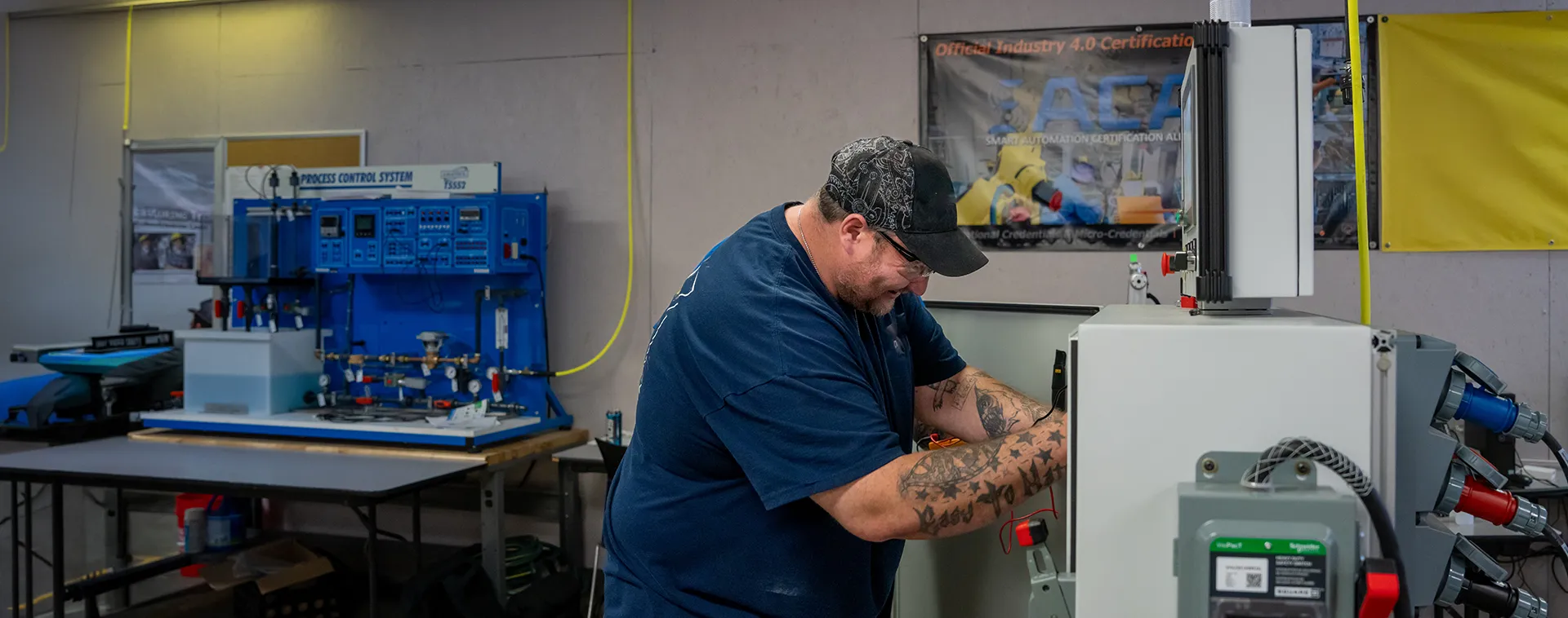 Person working on electrical panel in lab; blue process control system on table behind