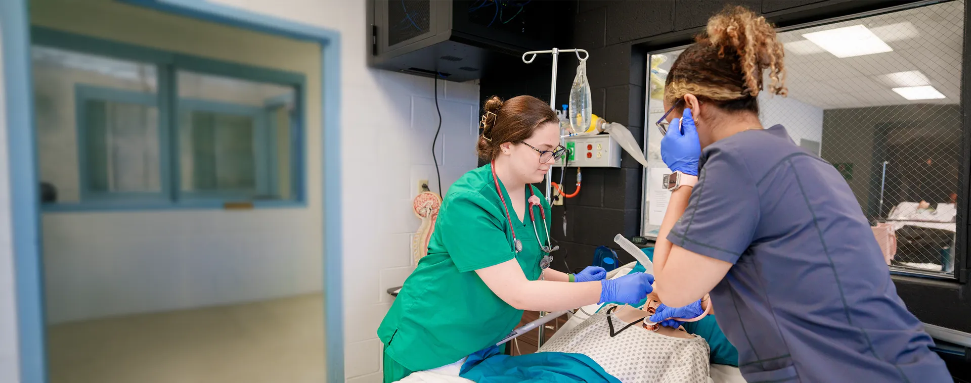Two students in scrubs practice a medical procedure on a training mannequin in a clinical lab with monitors and equipment