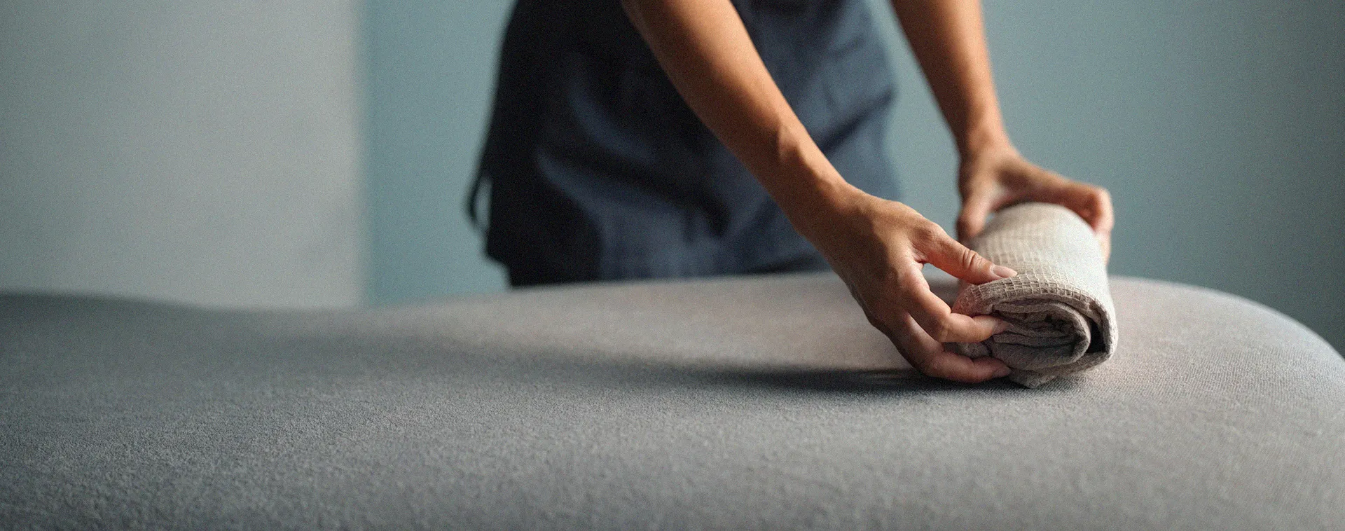 Hands rolling a folded towel on a massage table with gray fabric surface
