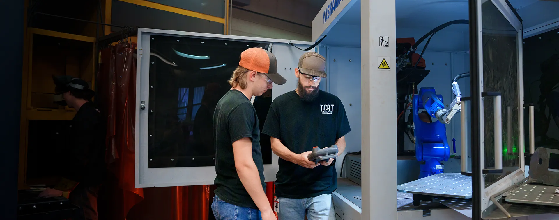 Two people wearing safety glasses review a handheld device beside a blue robotic arm inside an enclosed industrial lab