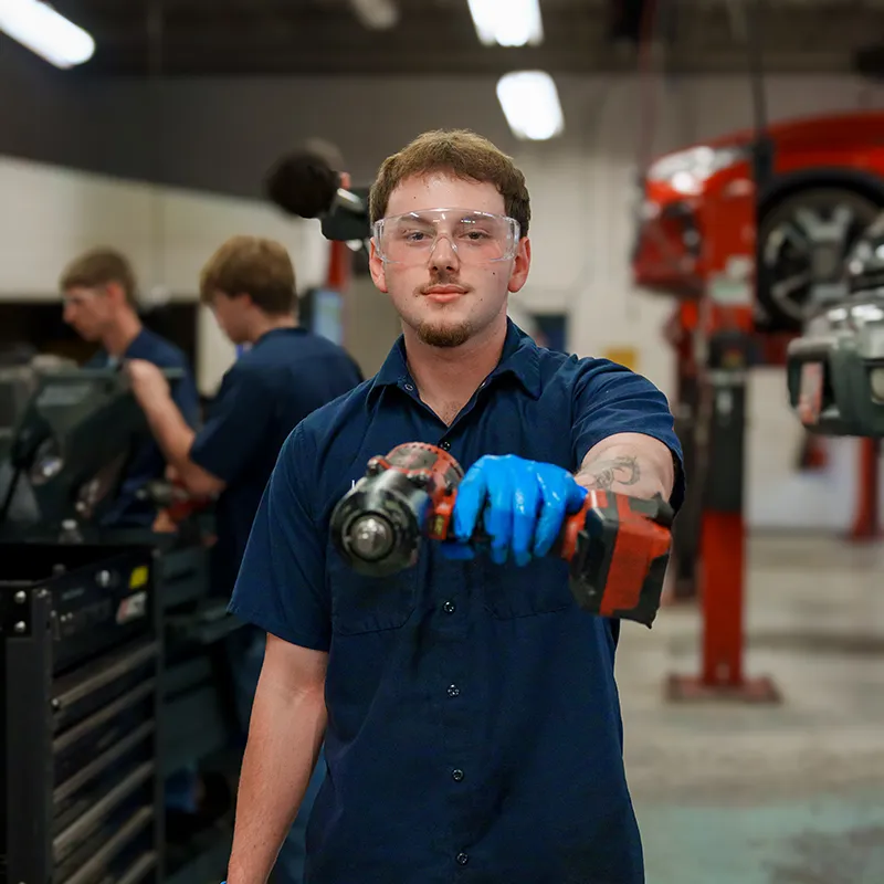 Student wearing safety glasses and gloves holding a power drill toward camera in automotive workshop