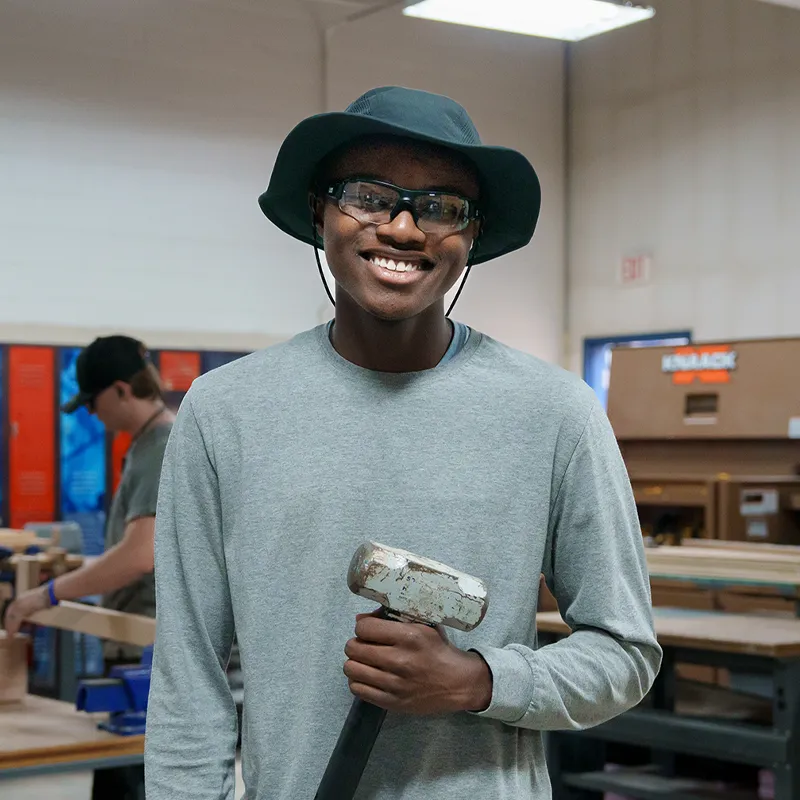 Student wearing safety glasses and hat holding mallet in workshop with tools and equipment in background
