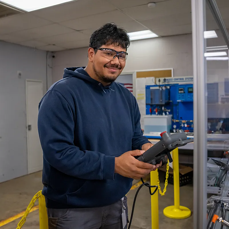 Student wearing safety glasses holding handheld control beside industrial equipment in workshop with yellow barriers