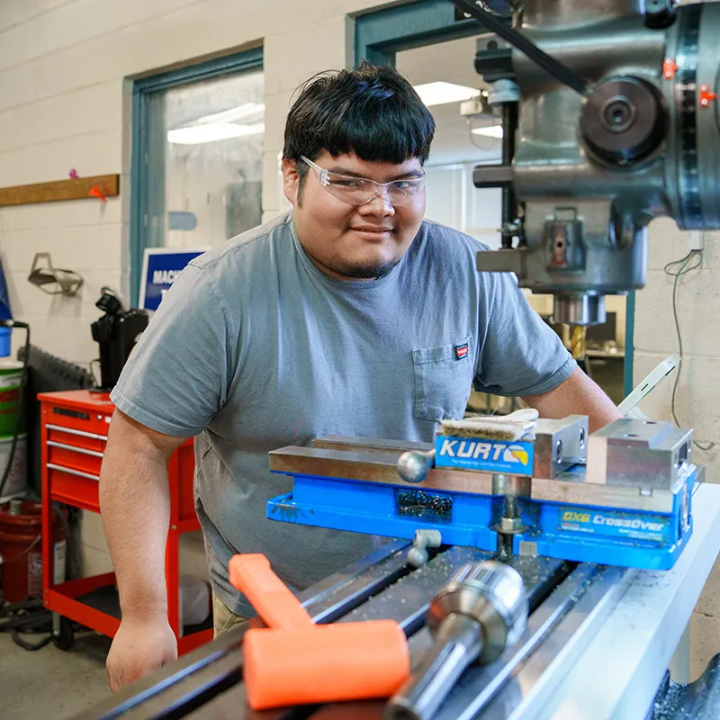 Student wearing safety glasses beside milling machine with vise and tools visible