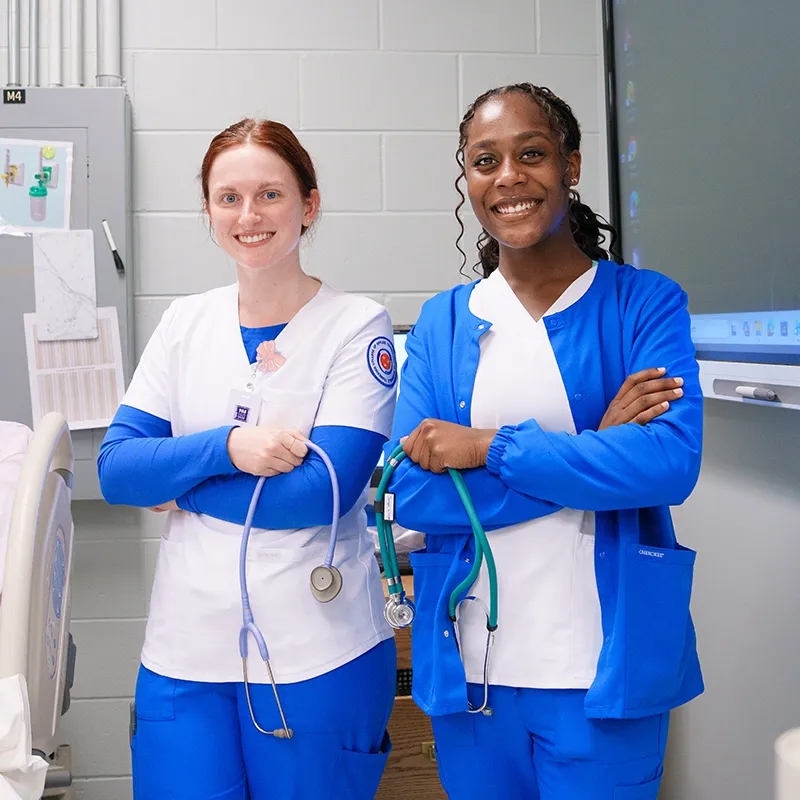 Two students in scrubs stand with arms crossed holding stethoscopes in a classroom with hospital bed and equipment