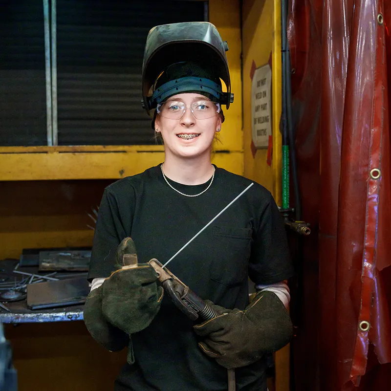 Student wearing welding helmet and gloves holds welding torch and filler rod in workshop with tools and red curtain backdrop