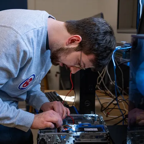 Student leaning over open desktop computer, connecting wires inside case with tools on table beside monitors