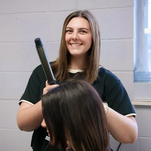 Student holding curling iron while working on another person&rsquo;s hair in classroom with window and light gray wall