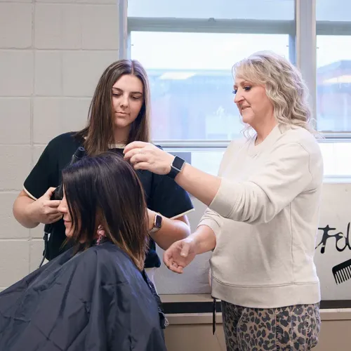 Instructor points to hair section while student uses curling iron on seated client near window with styling chair and cape