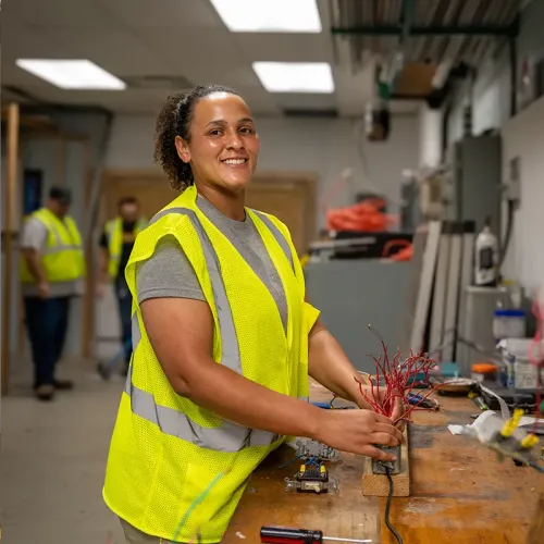 Person in safety vest works with exposed wiring on a table in electrical lab with tools and equipment visible in background