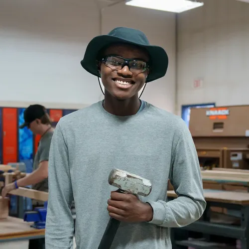 Student wearing safety glasses and hat holding mallet in workshop with tools and equipment in background
