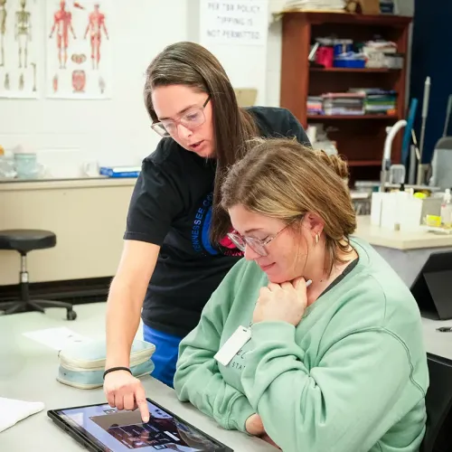 Instructor pointing at tablet screen while student looks on at table with anatomy charts and classroom supplies in background