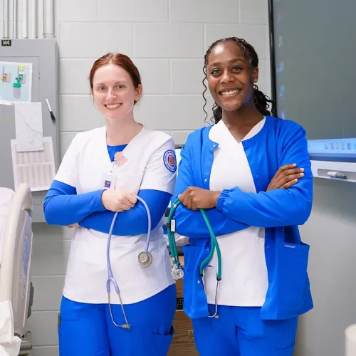 Two students in scrubs stand with arms crossed holding stethoscopes in a classroom with hospital bed and equipment