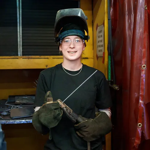 Student wearing welding helmet and gloves holds welding torch and filler rod in workshop with tools and red curtain backdrop