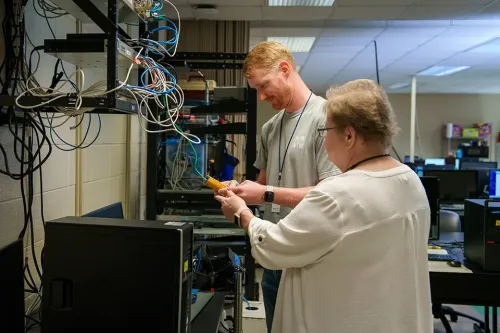 Two people working with network rack and cables, holding a yellow tool in a computer lab with equipment shelves