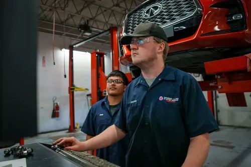 Two students in auto lab operating diagnostic computer near a raised red vehicle on a lift
