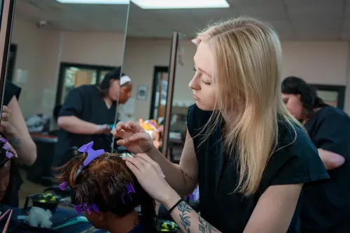 Student pins rollers into mannequin head at workstation; mirrors, tools, and other students visible in background