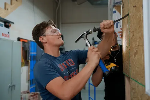 Student hammering nail into wall panel while another holds power drill behind in lab