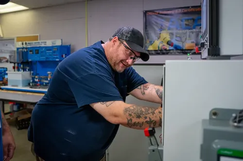 Person with tattooed arms works inside electrical control cabinet in workshop with equipment and certification poster