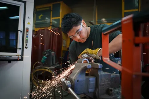 Student uses a handheld grinder on metal clamped in a vise, sparks flying in a workshop with equipment and safety curtains