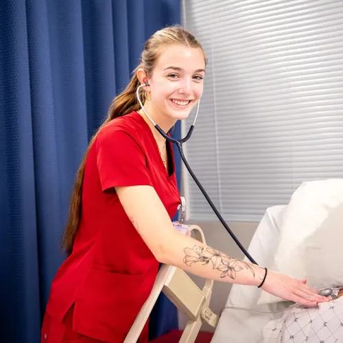 Student in red scrubs using a stethoscope on a patient bed in a healthcare training lab.