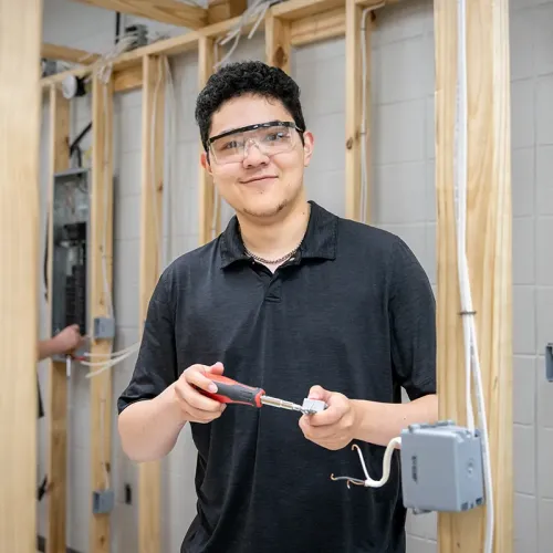 Student wearing safety glasses using a screwdriver to wire an electrical box in a framed training wall.